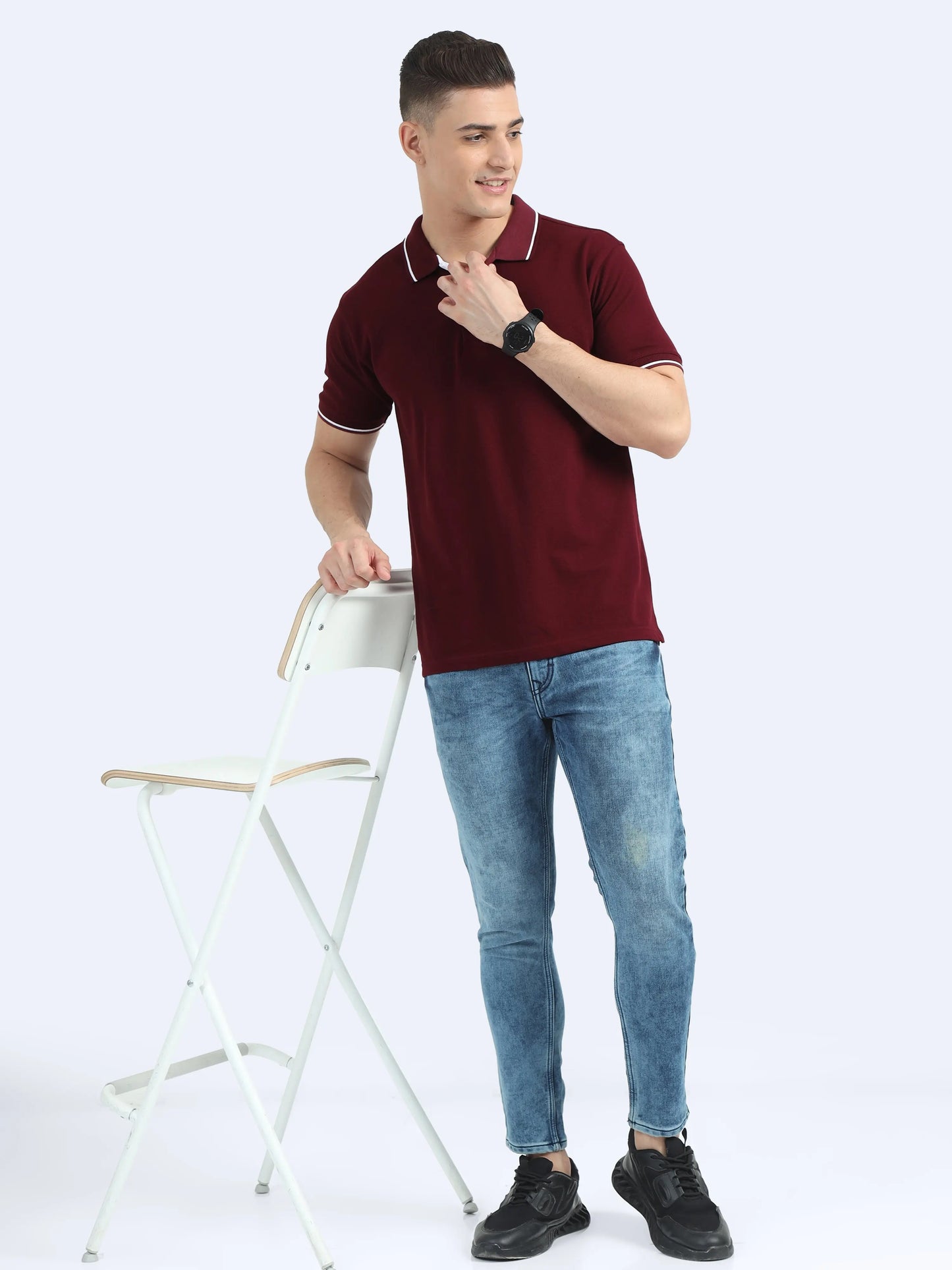 Man wearing maroon polo t-shirt and blue jeans standing by a white chair, casual apparel