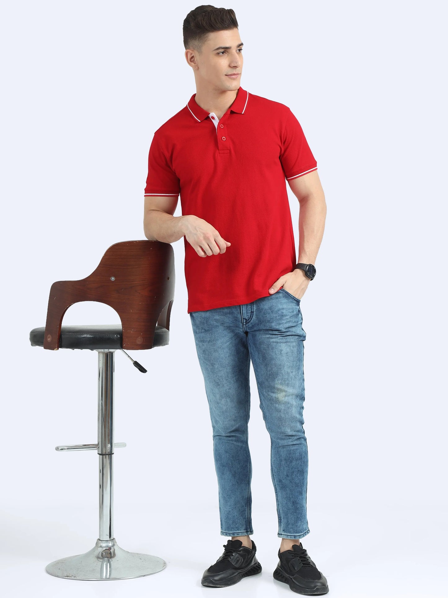 Man in red polo t-shirt and jeans posing beside a modern wooden bar stool, casual fashion.