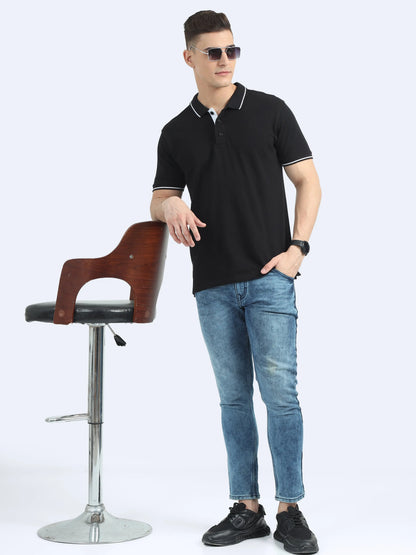 Man wearing black polo shirt, blue jeans, and sunglasses, standing by a modern wooden chair