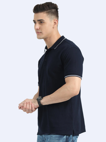 Man wearing navy blue polo t-shirt with white trim and jeans, posing against light background
