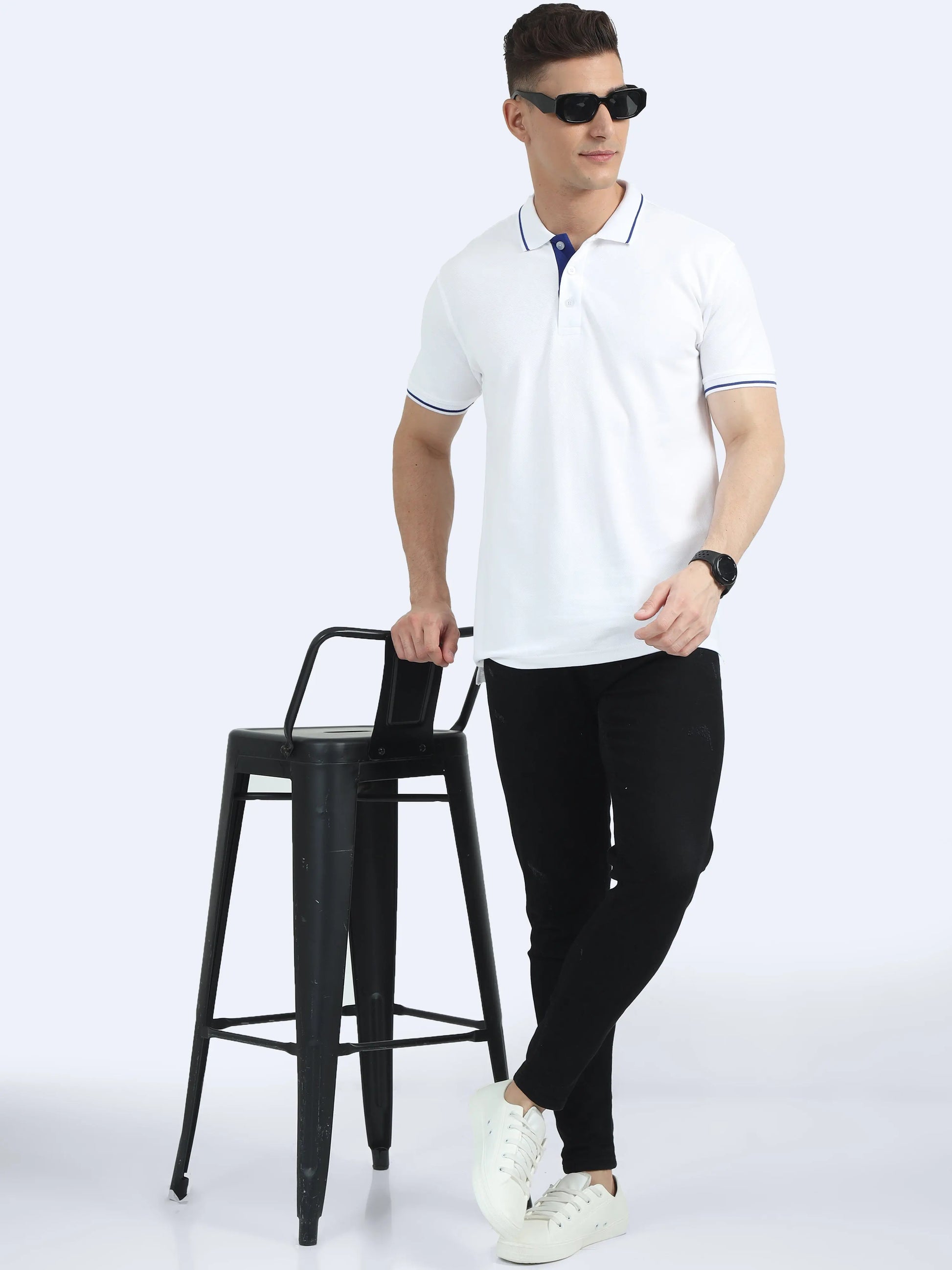 Man in white polo shirt and black pants with white sneakers standing by black stool