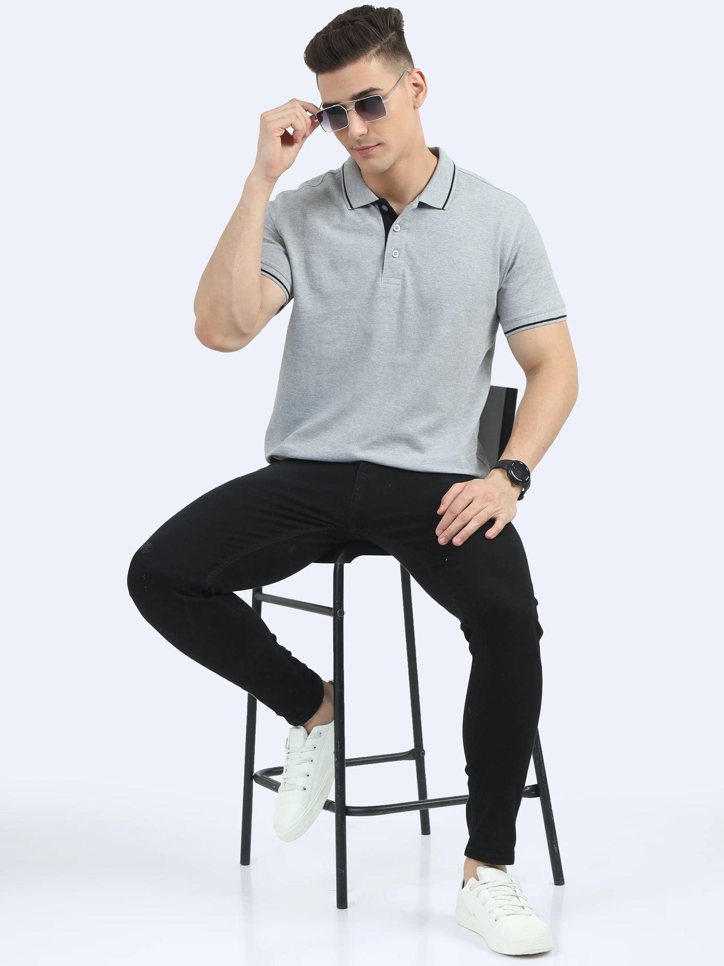 Man in grey polo shirt and black pants seated on a stool, wearing sunglasses and white sneakers