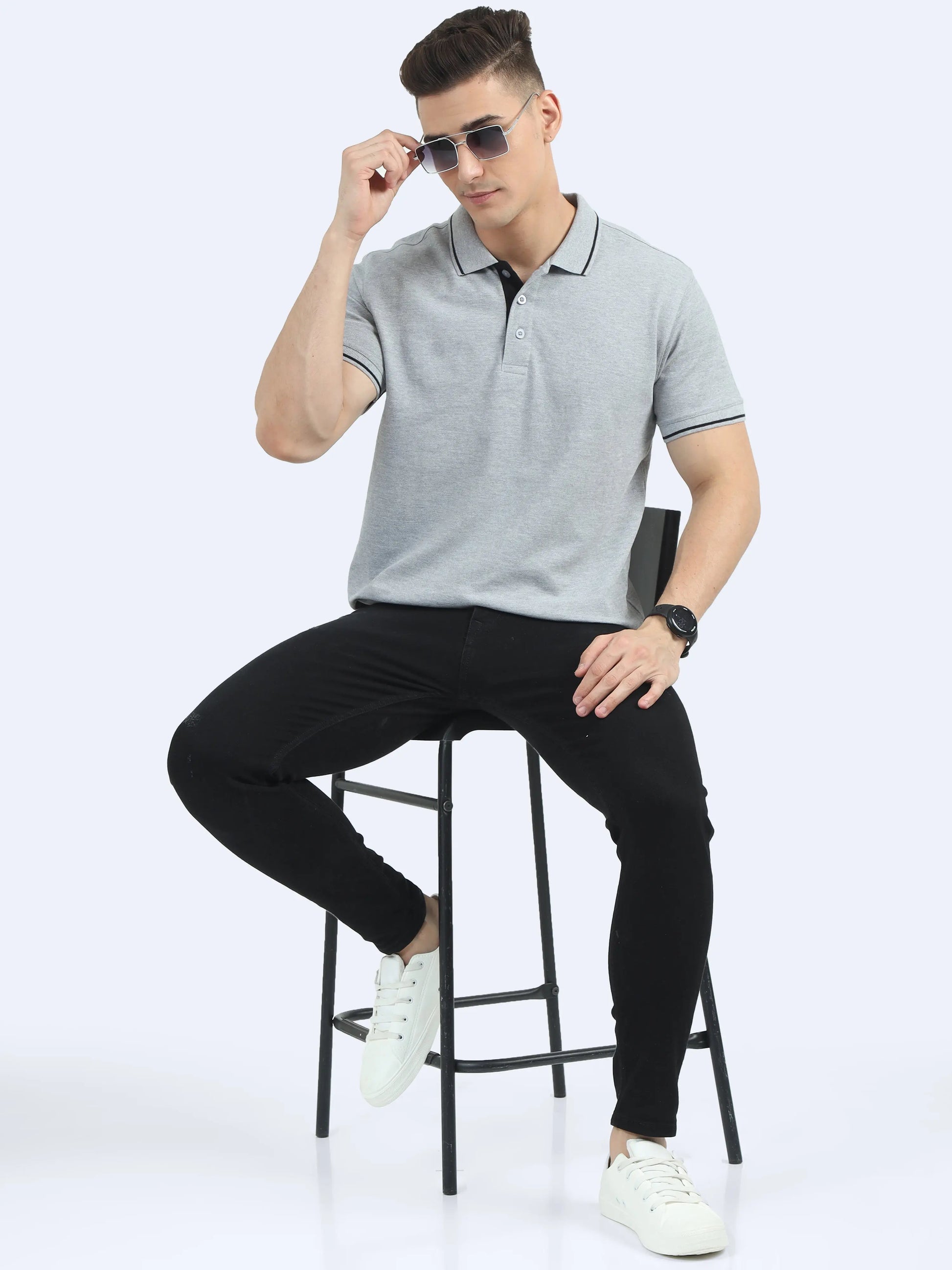 Man in grey polo shirt and black pants seated on a stool, wearing sunglasses and white sneakers