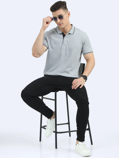 Man in grey polo shirt and black pants seated on a stool, wearing sunglasses and white sneakers