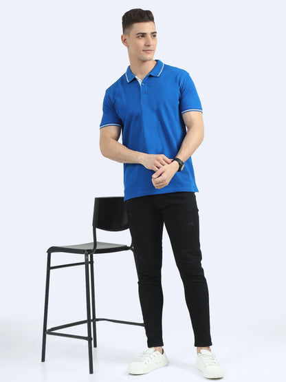 Man wearing royal blue polo shirt and black jeans, standing next to bar stool on white background