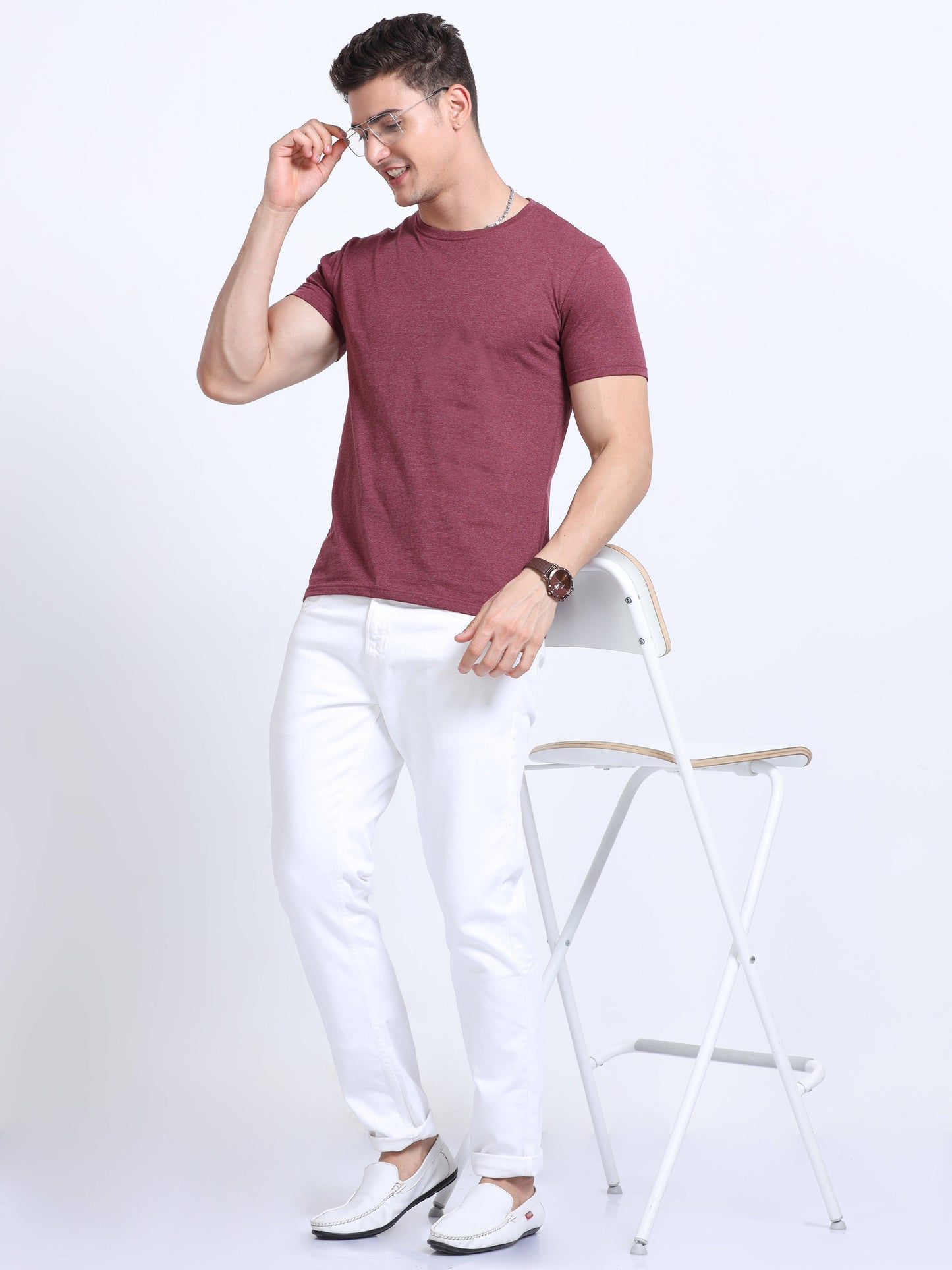 Man in maroon t-shirt and white pants posing with glasses next to a white chair
